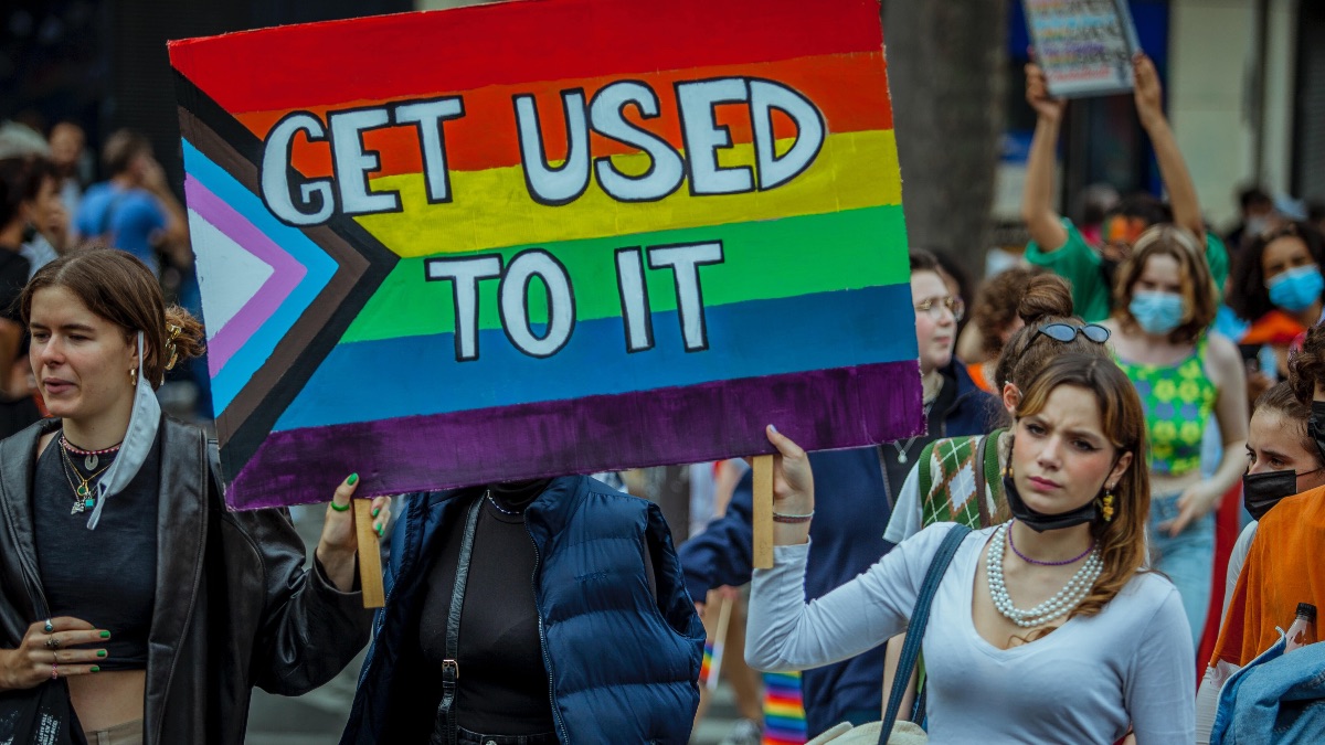 Two people at protest hold placard painted with trans inclusive pride flag, writing reads 